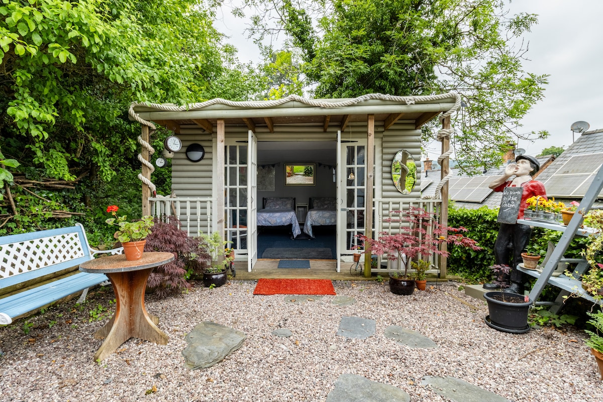 The cabin, nestled within a garden, features a welcoming entrance with a red mat. A small table and chairs are positioned outside, surrounded by potted plants and gravel. Inside, two single beds are visible through the open glass doors, inviting relaxation.