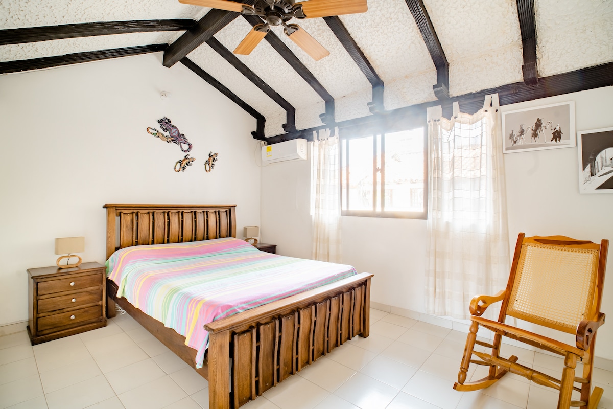 A spacious room is presented with a wooden bed adorned in a multicolored striped blanket. Natural light filters through the window, illuminating the space. A ceiling fan and air conditioning offer comfort, while a rocking chair adds a decorative touch.