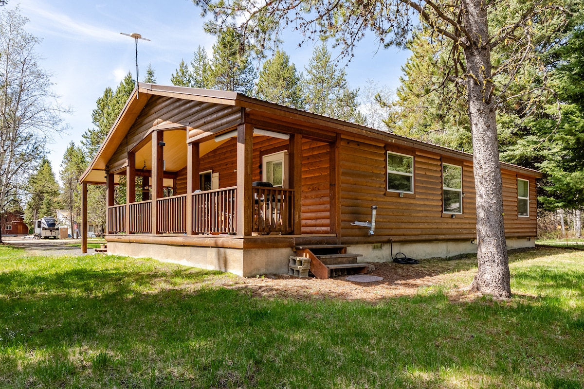 A single-story cabin showcases a wooden exterior with a covered porch and railings. The surrounding green lawn is bordered by trees, providing a natural setting. Windows allow for ample natural light, emphasizing the structure's inviting appearance.