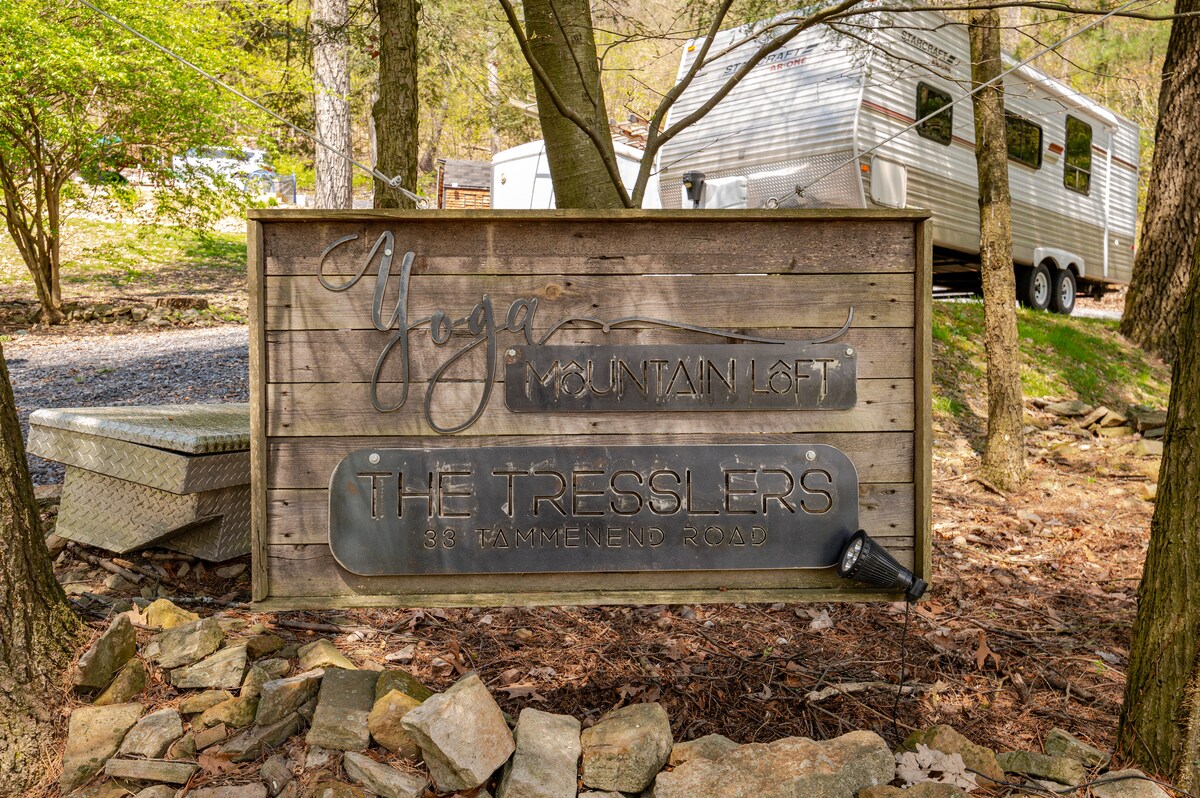 A wooden sign marks the entrance to Yoga Mountain Loft, featuring the name 'The Tresslers' prominently displayed. The background includes a travel trailer parked among trees, with a gravel driveway visible and natural stone accents surrounding the sign.