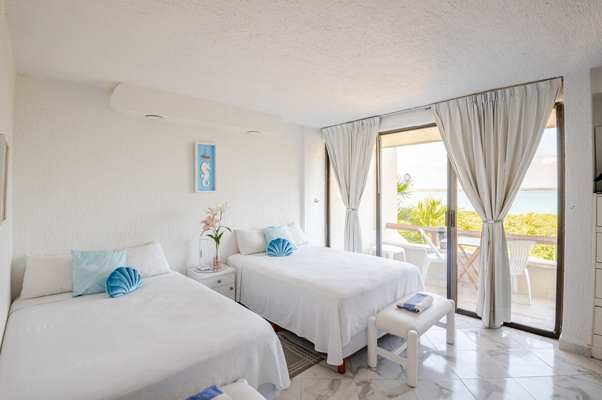 The studio features two spacious beds dressed in white linens, complemented by decorative cushions. Large glass doors open to a balcony with a table and chairs, offering views of the lagoon. Light-coloured curtains frame the windows, enhancing the airy atmosphere.