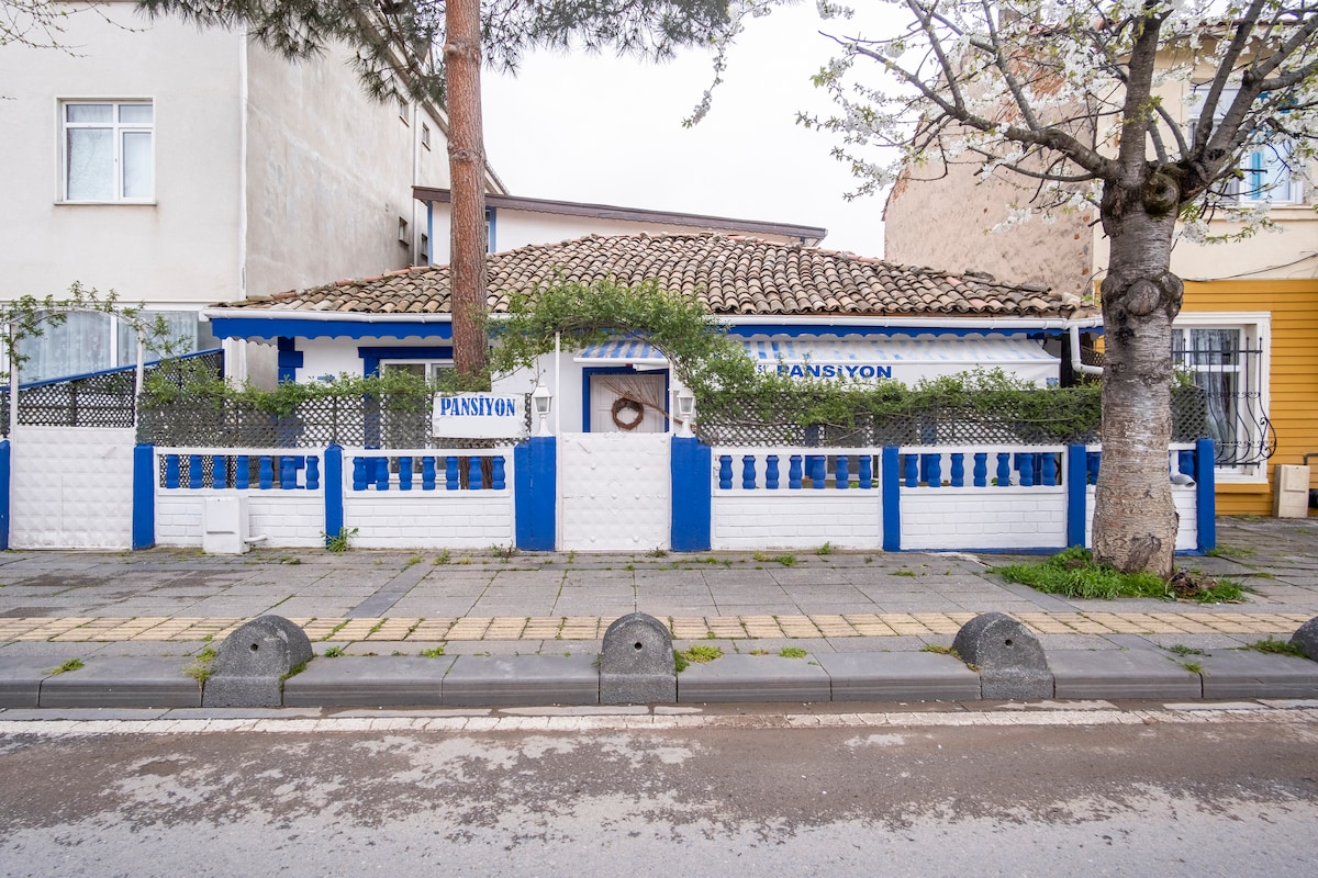 The exterior of a detached house is shown, featuring a tiled roof, blue accents, and white walls. A fence surrounds the property, and a small porch area is visible. A tree with bare branches stands in the front yard, providing some greenery.