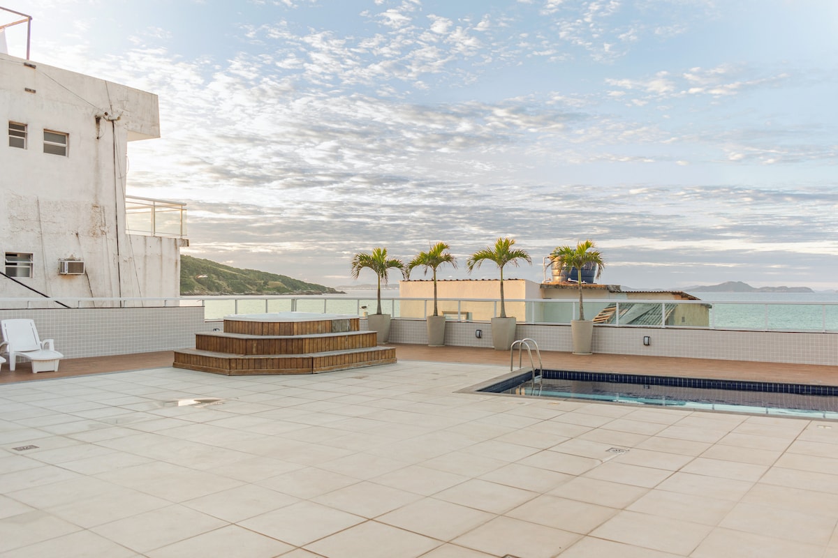 A spacious rooftop terrace is presented, featuring a rectangular pool and seating area made of wooden deck. Palm trees can be seen in the background, along with a view of the ocean and distant hills. The sky displays soft clouds during a tranquil evening light.