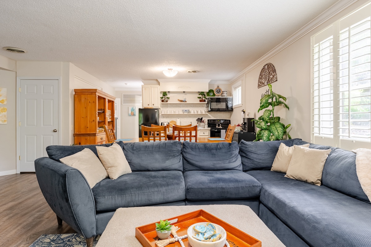 A spacious living area features a large sectional sofa in soft blue, with decorative cushions. A light-colored coffee table is centered in the room, adorned with a tray holding decorative items. The open layout connects to a bright kitchen, visible in the background.