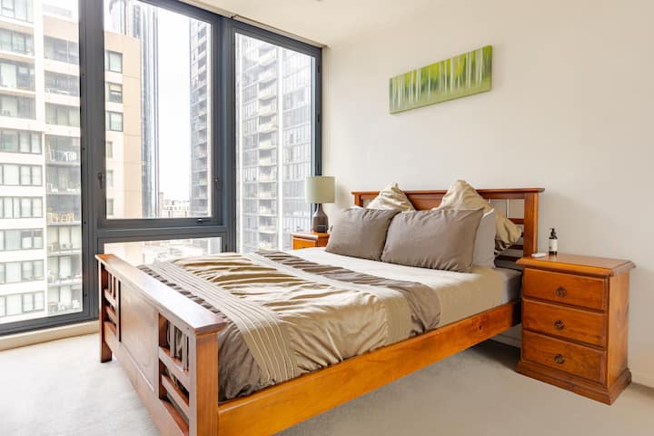 A modern bedroom with large floor-to-ceiling windows overlooking nearby high-rise buildings. The room features a wooden bed frame with beige bedding and matching pillows, flanked by two wooden bedside tables. A minimalist painting hangs above the bed, adding a touch of color to the space.