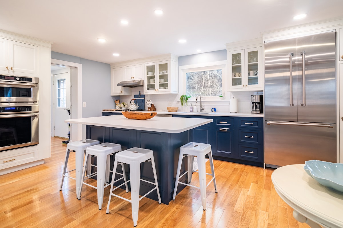 A modern kitchen features a large island with three metal stools, complemented by white cabinetry and blue accents. Stainless steel appliances include an oven and a refrigerator. Natural light illuminates the space through a window above the sink, providing a welcoming and functional atmosphere.