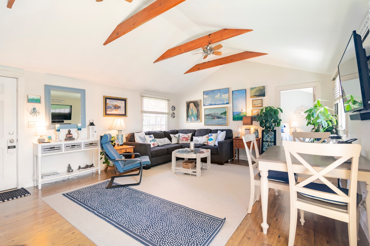 An airy living space features vaulted ceilings with wooden beams, a large sectional sofa adorned with colorful cushions, and a light-colored rug. A dining area with a wooden table and chairs is situated beside a potted plant. Wall art depicting coastal scenes adds a vibrant touch.