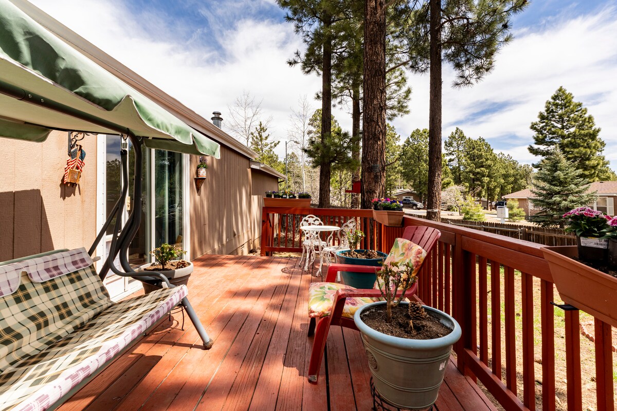 An outdoor deck features a glider and several potted plants, surrounded by trees. A small table and chairs offer a cozy space for relaxation. The wooden deck has a warm finish, complementing the natural landscape beyond.