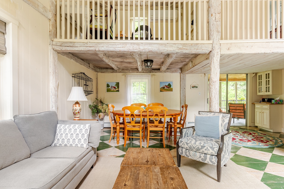 An inviting living space features exposed beams and a spacious layout. A comfortable sofa and accent chair provide seating, while a rustic coffee table anchors the area. A large farmhouse dining table with seating for six is positioned nearby, surrounded by artwork and natural light.