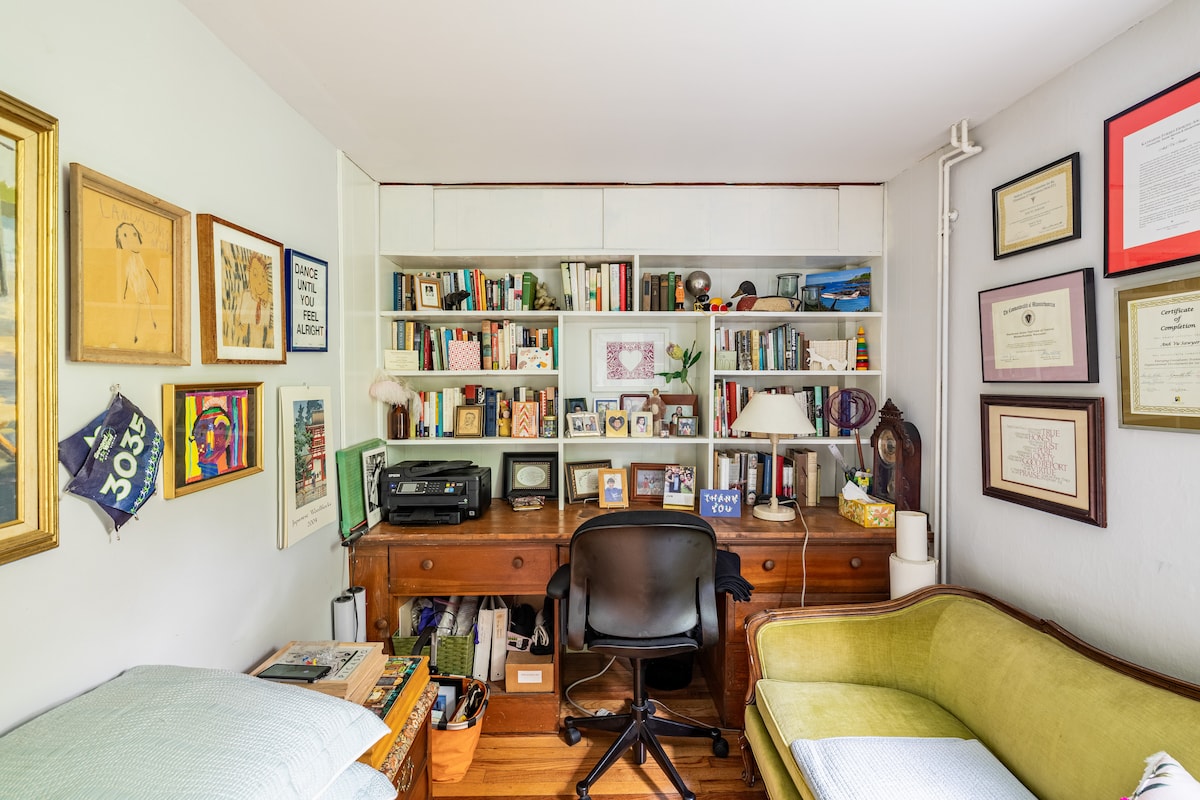 A small home office is featured, with a wooden desk positioned against a bookshelf filled with books and decorative items. Framed artwork and certificates adorn the walls. A green couch is situated alongside the desk, creating a cozy reading nook.