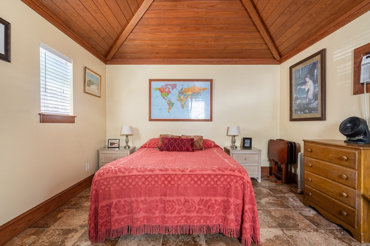 A cozy sleeping area features a spacious bed covered with a red patterned blanket. The tongue and groove wood ceiling adds warmth, while two bedside tables with lamps flank the bed. Light enters through windows, and a world map is displayed on the wall.
