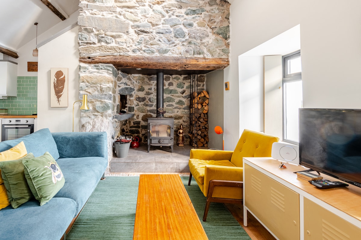 A comfortable living area displays a blue sofa and a yellow armchair, arranged around a wooden coffee table. The stone wall features a wood-burning stove and a neatly stacked pile of firewood. Natural light enters through a large window, enhancing the open-plan space.