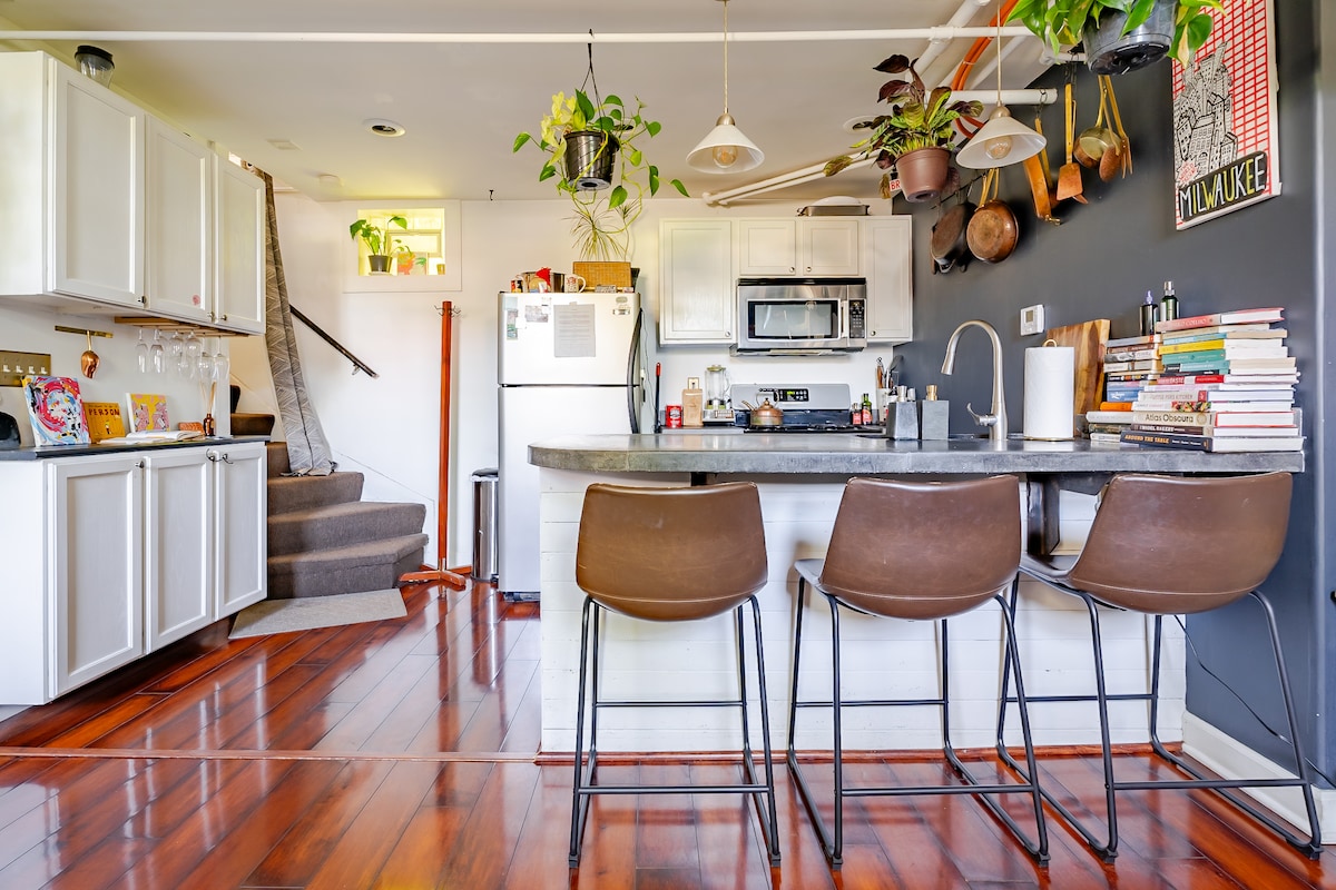 The open concept kitchen features a sleek countertop with three high-backed stools. Light wooden cabinets contrast with dark walls adorned with colorful artwork. A refrigerator and microwave are visible, complemented by plants hanging above the space, while a staircase leads to the upper level.