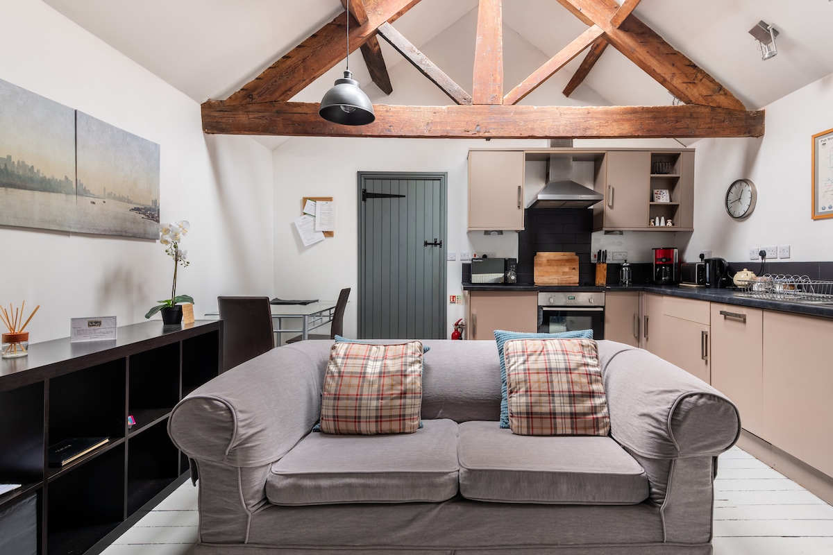 The vaulted lounge area features exposed wooden beams and a comfortable settee with plaid cushions. A modern kitchen with quality appliances is visible in the background, alongside a dining table and chairs. Natural light illuminates the space from a nearby window.