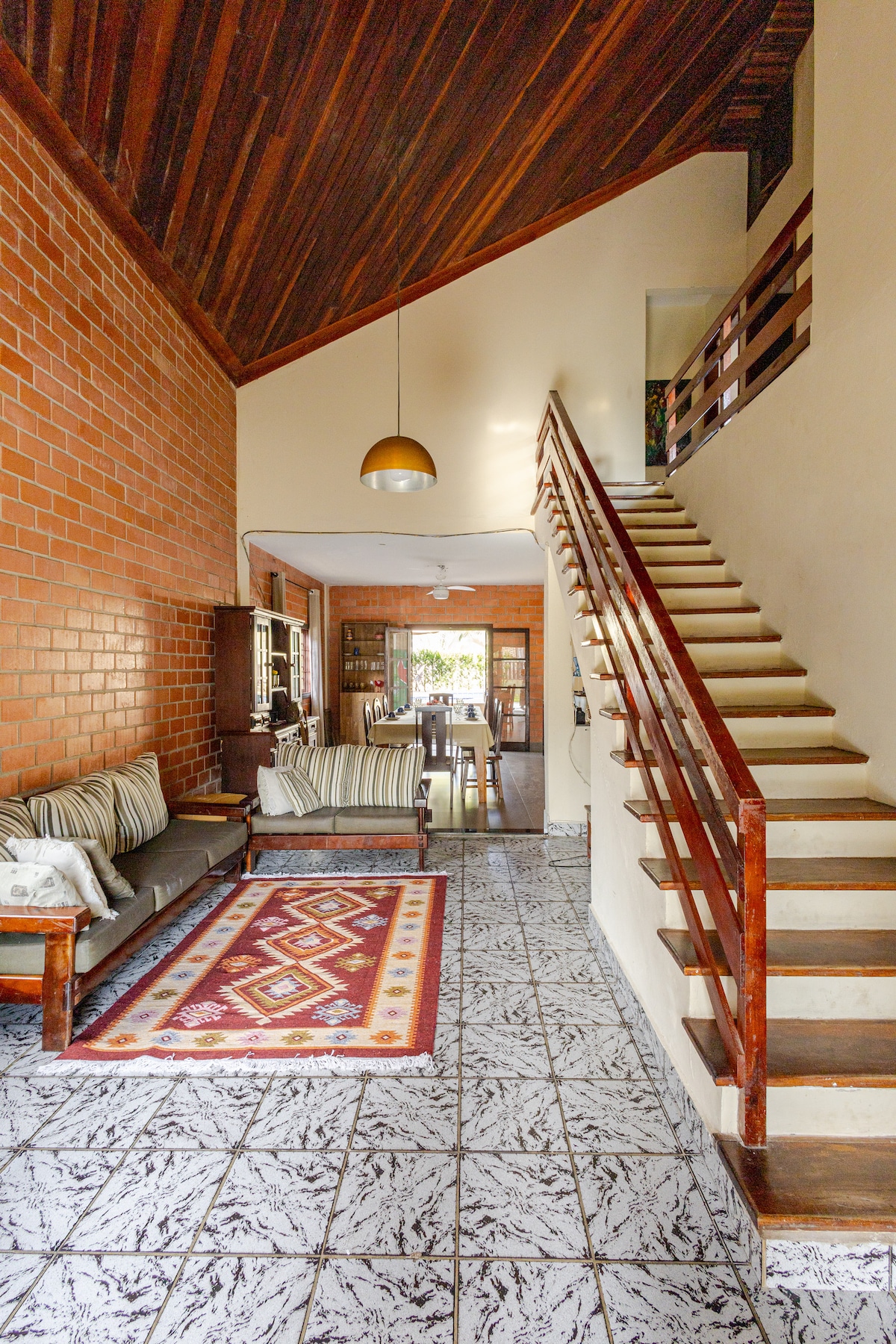 An inviting common area features high wooden ceilings and exposed brick walls. A cozy seating arrangement with striped cushions is placed on a patterned rug. Stairs lead to an upper level, while dining space is visible through large windows, allowing natural light to fill the room.