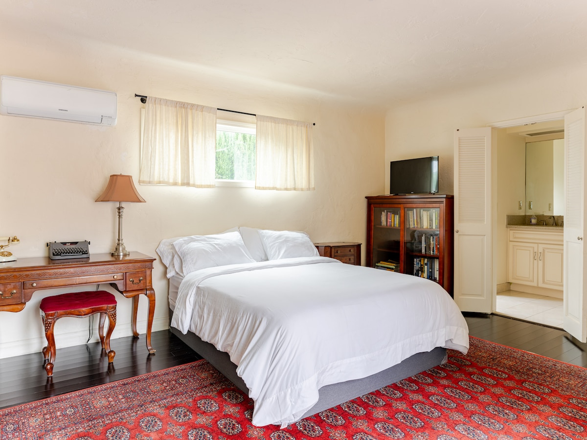 A bedroom features a spacious bed dressed in white linens, accompanied by a wooden desk and a lamp. Natural light filters through sheer curtains. A bookcase stands against the wall, and a television is positioned in an adjacent area, visible through an open doorway.
