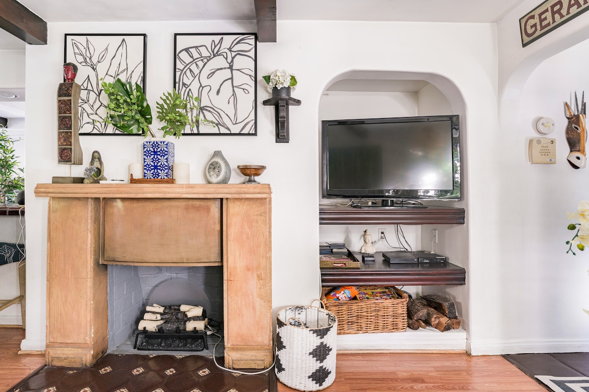 A cozy living area features a decorative fireplace with a minimalist design, complemented by framed botanical art on the walls. A television is neatly integrated into the shelving beneath, while a woven basket and a patterned storage container provide additional texture and functionality.
