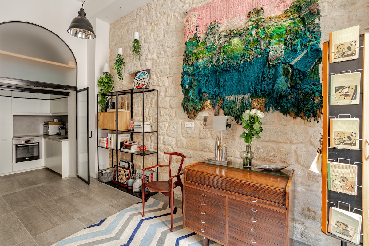 An open living space features textured stone walls and a large, colorful wall hanging. A mid-century style wooden console table sits alongside a vintage chair. A modern kitchen is visible in the background, equipped with sleek cabinetry and appliances, while a striped rug adds a subtle design element.