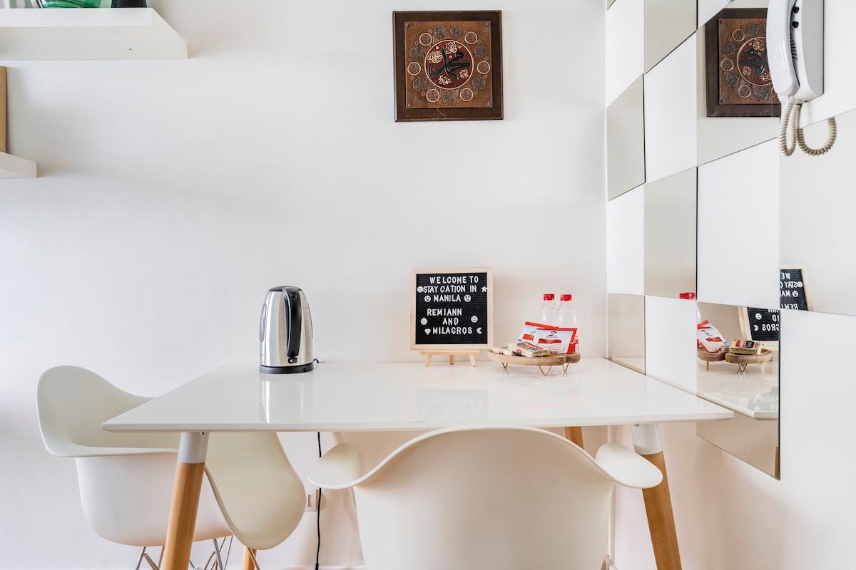 A bright dining area features a sleek white table with wooden legs and four matching chairs. A kettle is placed on the table, alongside a small sign welcoming guests. Reflective surfaces create an airy feeling, while wall decor adds a touch of character.