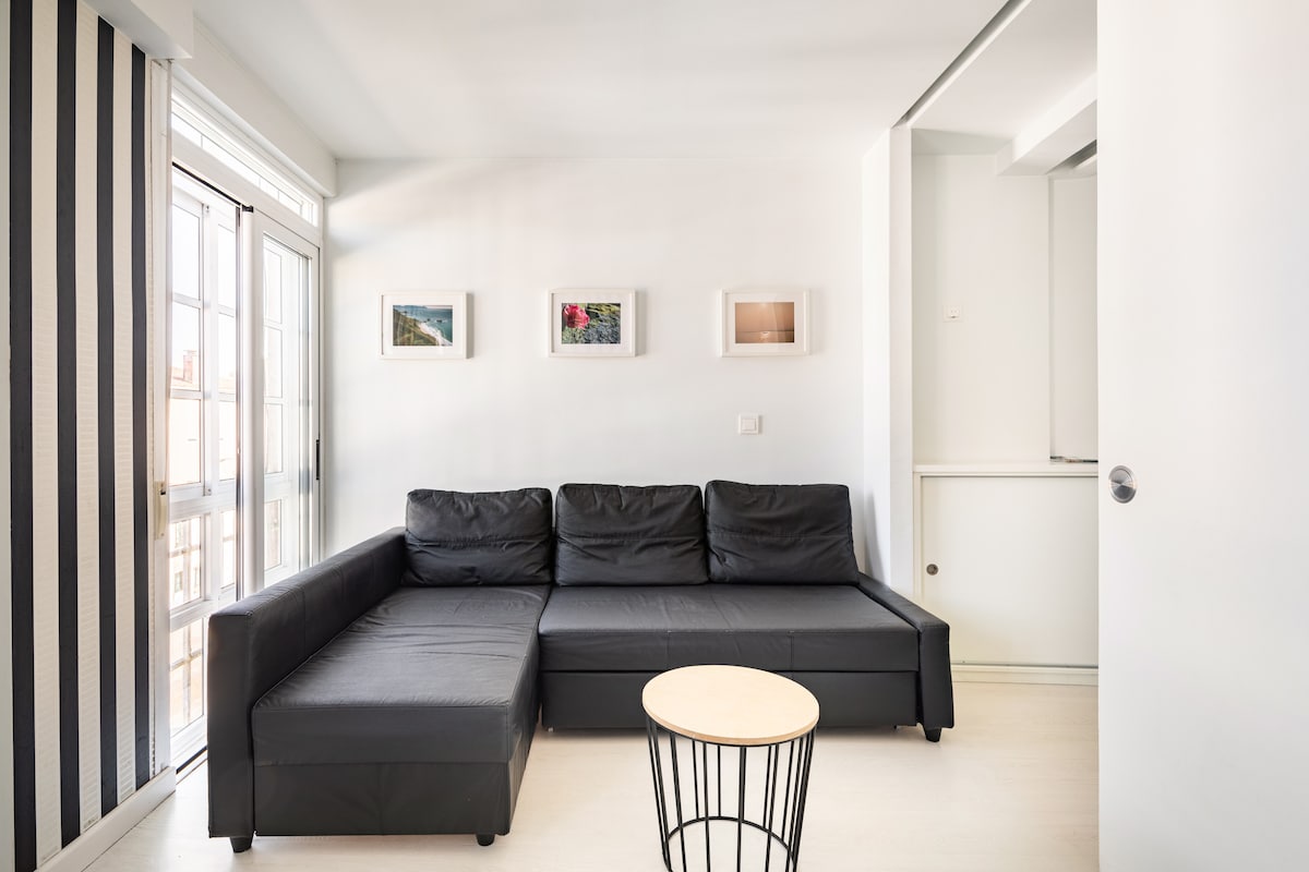 A modern living space features a black sectional sofa alongside a small round coffee table. Framed pictures adorn the white walls, while large windows provide natural light, with sheer curtains allowing for privacy. The area is designed for comfort and relaxation.