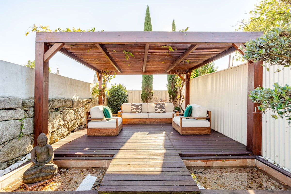 A gazebo offers a serene seating area, featuring two cushioned sofas on a wooden deck. A Buddha statuette is positioned peacefully on the ground, surrounded by gravel and bordered by vertical wooden fencing and lush greenery.