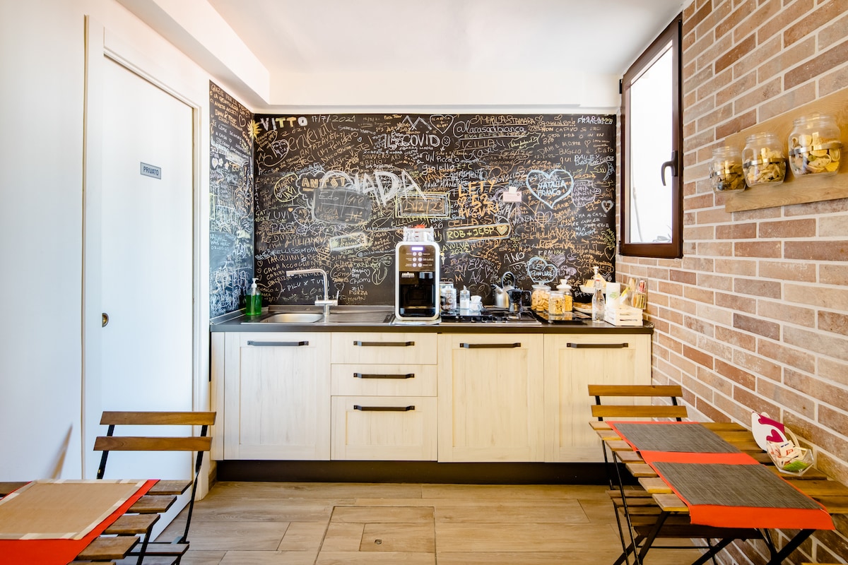A communal kitchen area is equipped with modern cabinetry and a coffee machine. One wall is adorned with chalkboard surfaces displaying various doodles and notes. Seating is provided by two small tables with chairs, contributing to a welcoming environment for shared meals.