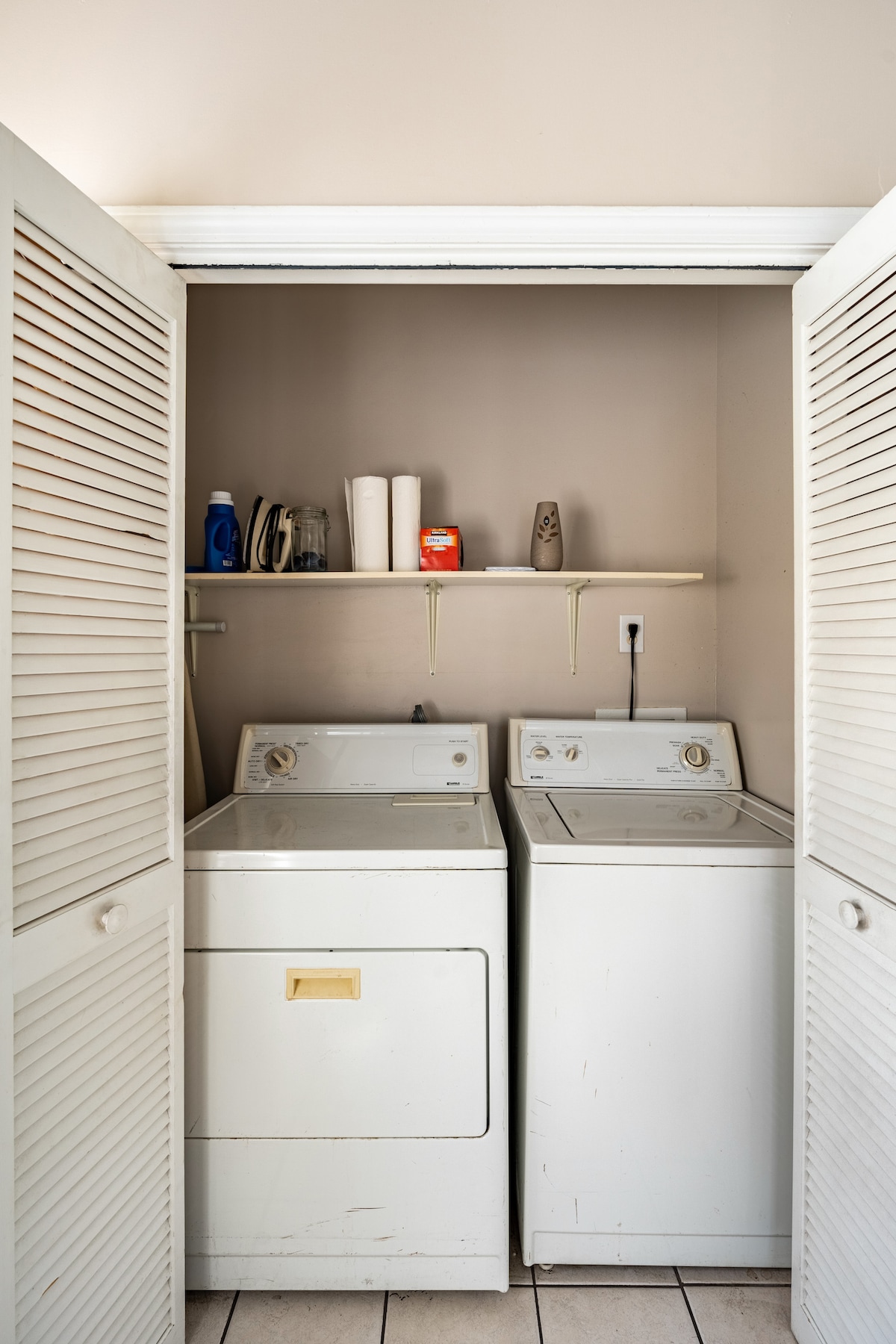 A laundry area is presented with a stacked washer and dryer, placed side by side. Above, a shelf holds various laundry supplies. The space is enclosed by folding doors, providing a functional utility within the apartment.