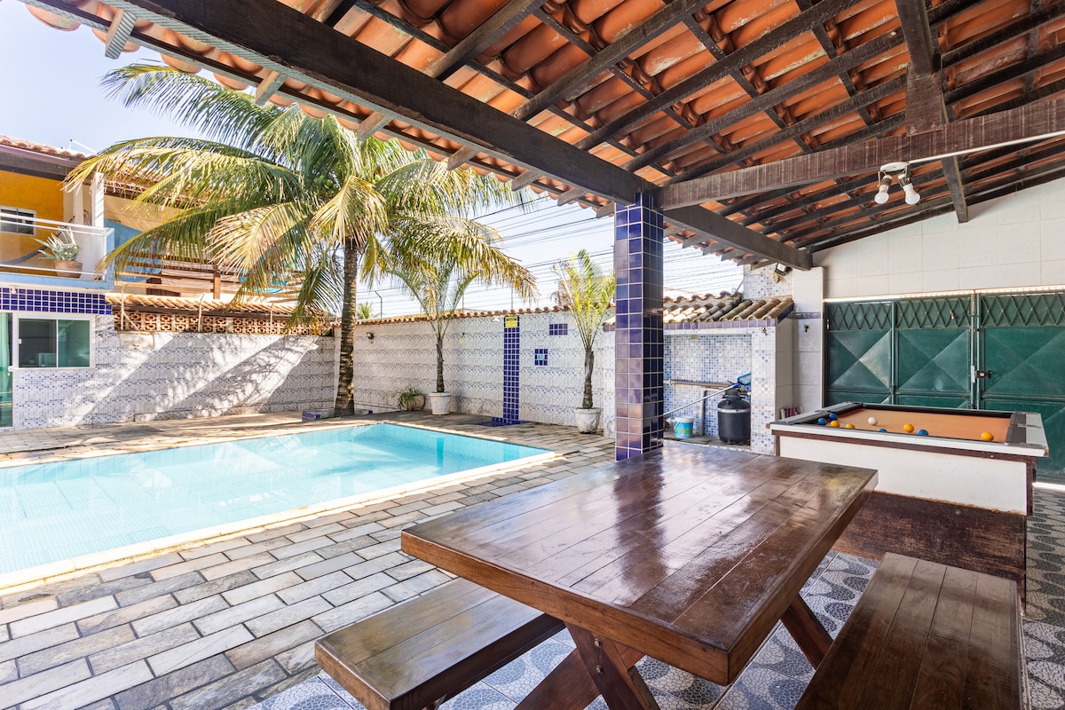 An outdoor area is shown, featuring a private swimming pool alongside a wooden table. A billiard table is visible, with various palm trees providing shade. The surrounding walls are decorated with tiles, and the roof is made of terracotta tiles, creating a cozy atmosphere.