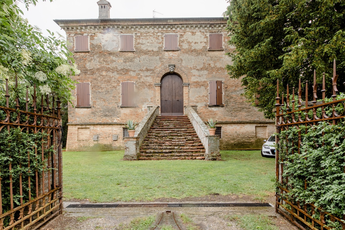 The villa's elegant façade features weathered brick walls and large wooden doors, flanked by a set of stairs. Lush greenery surrounds the entrance, and an ornate wrought-iron gate leads into the spacious lawn, providing a sense of warmth and charm.
