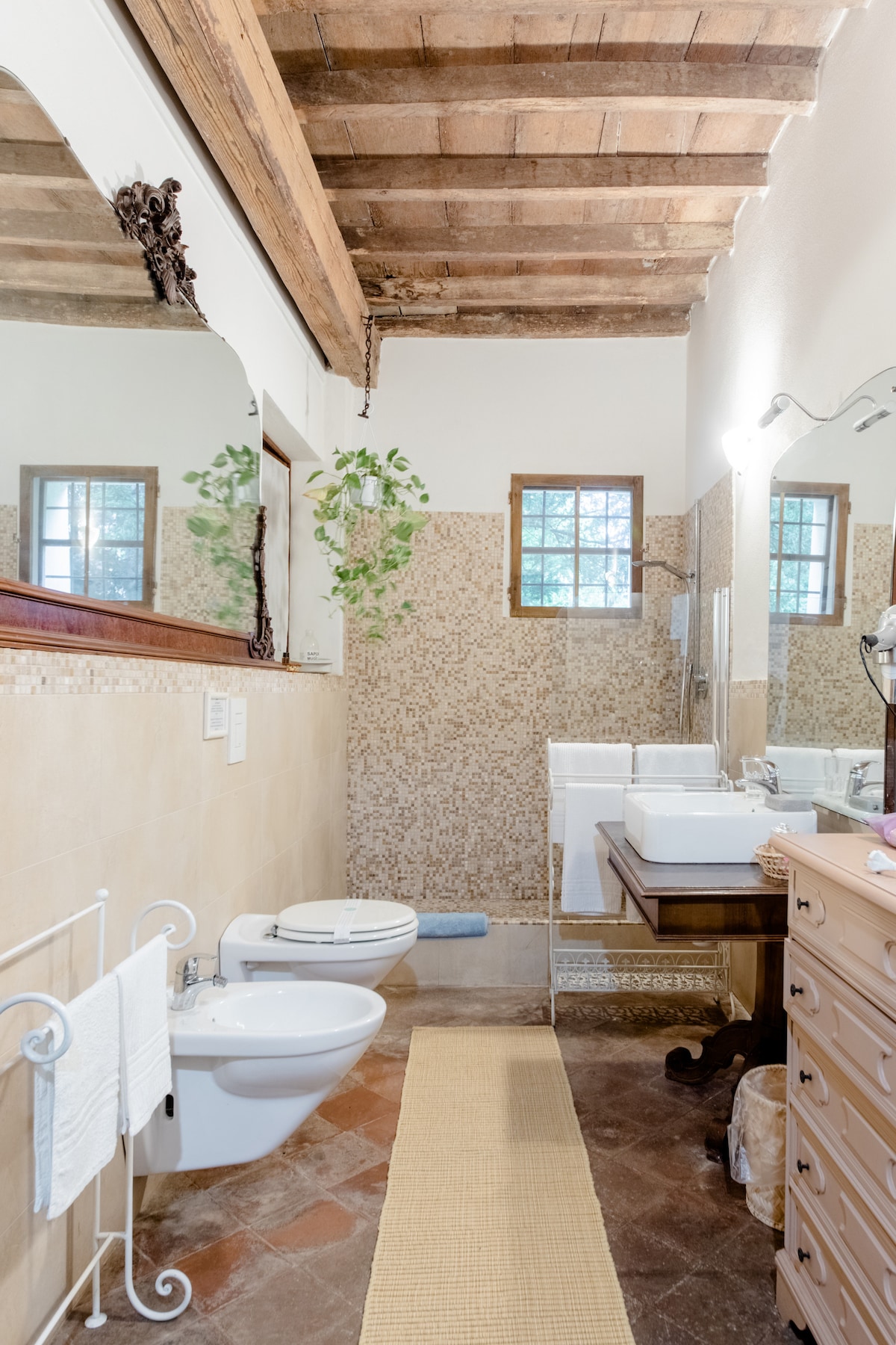The bathroom features warm-toned tiles and a wooden ceiling with exposed beams. A spacious sink is paired with a wooden vanity, while two bidets are positioned next to each other. Natural light filters in through the windows, enhancing the serene ambiance.
