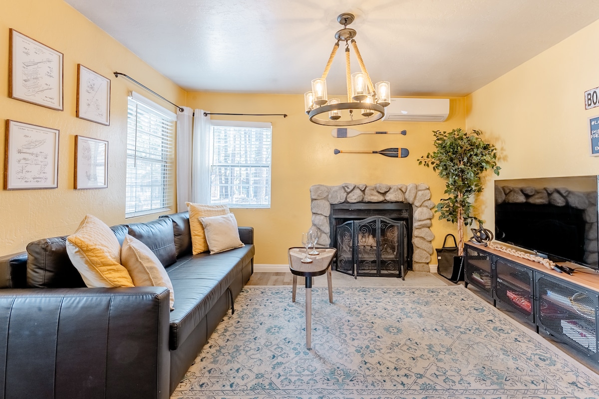 A welcoming living area features a large leather sofa paired with a round coffee table. A stone fireplace adds warmth to the space, complemented by a chandelier overhead. Natural light enters through multiple windows, with an indoor plant enhancing the cozy atmosphere.