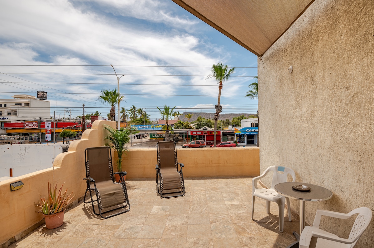 The terrace features two reclining chairs and a small table, offering a comfortable space for relaxation. Surrounding palm trees and a view of the nearby street enhance the outdoor experience, while bright skies create an inviting atmosphere.
