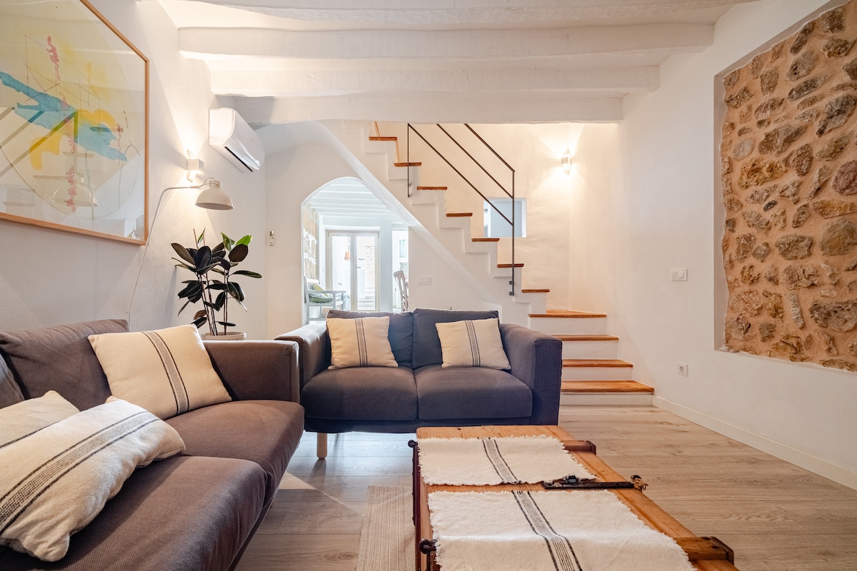 A comfortably furnished living area features two cozy sofas adorned with cushions. A wooden coffee table adds a natural element. The room is illuminated by soft lighting, with an artistic wall hanging and a potted plant contributing to the inviting environment. A staircase leads to the upper level.