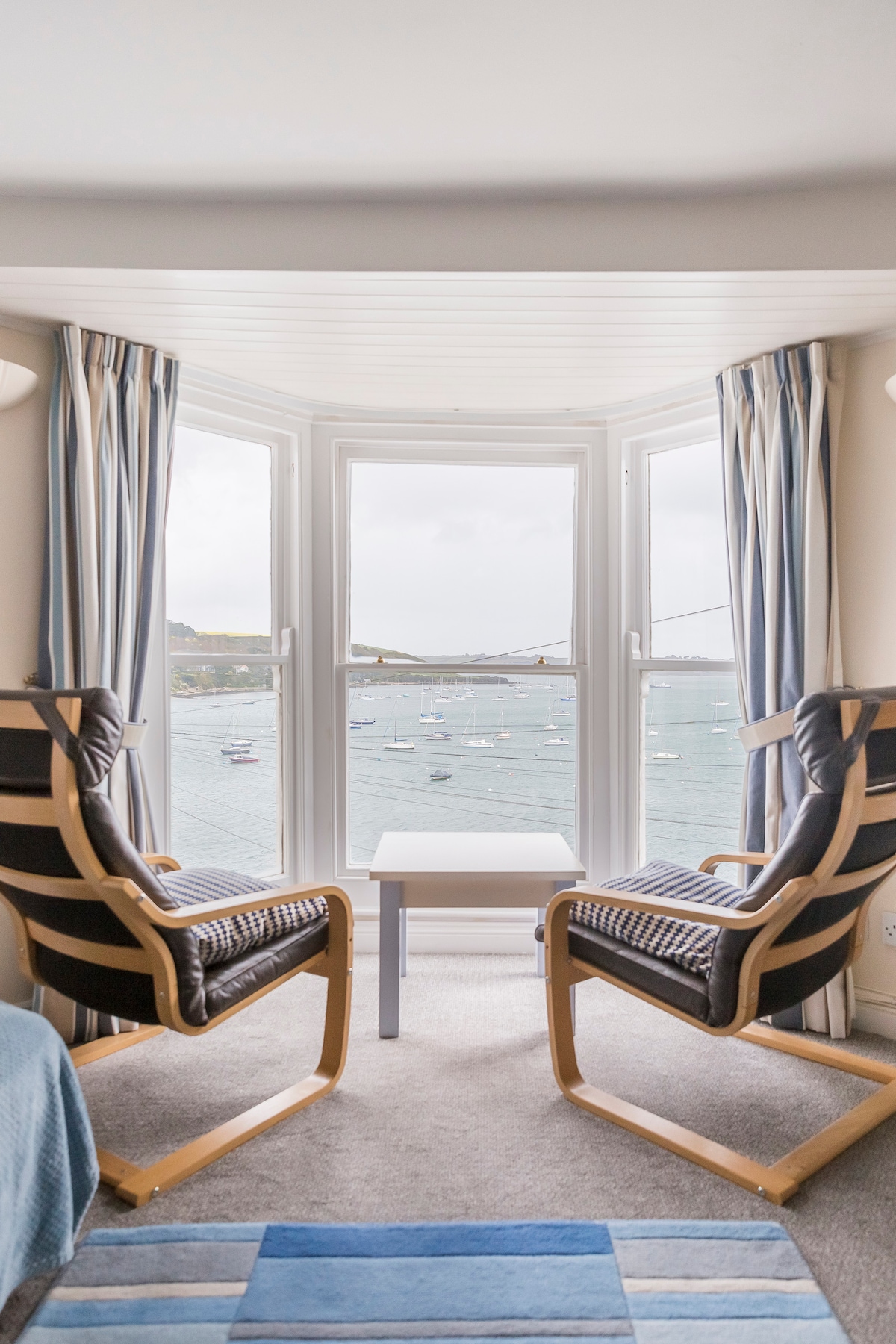 The image captures a bright seating area featuring two comfortable armchairs with patterned upholstery. A small table stands between them, with large windows framing a scenic view of the harbor and boats on the water. Soft curtains in shades of blue and gray complement the decor.