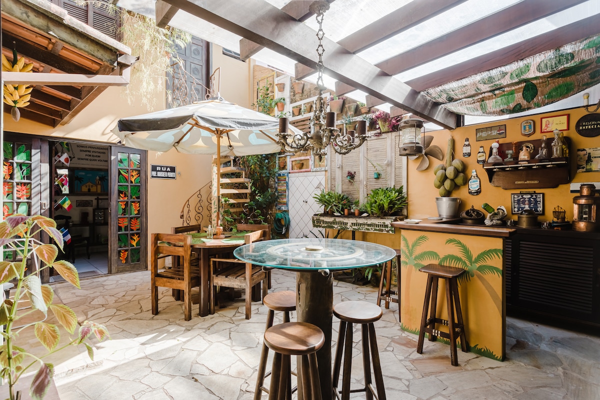 A lively communal kitchen and dining area is illuminated by natural light through the glass roof. Wooden tables and stools are arranged around a circular glass table. A charming spiral staircase leads to an upper level, and decorative plants enhance the inviting ambiance.