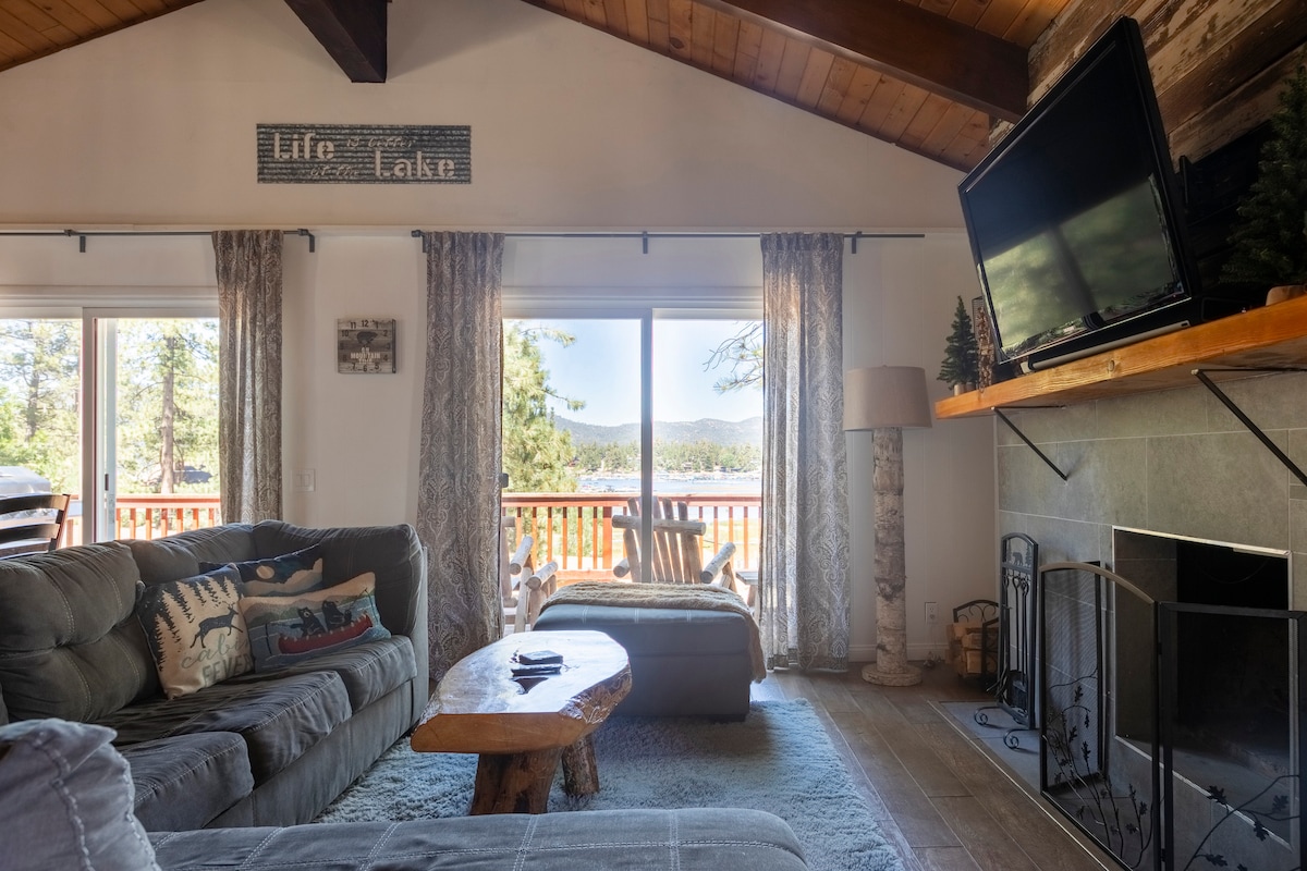 A spacious living area features vaulted wooden ceilings and large windows offering views of the lake and mountains. Comfortable seating is arranged around a natural wood coffee table, with a stone fireplace adding a cozy touch. Soft, neutral tones create a calming environment.