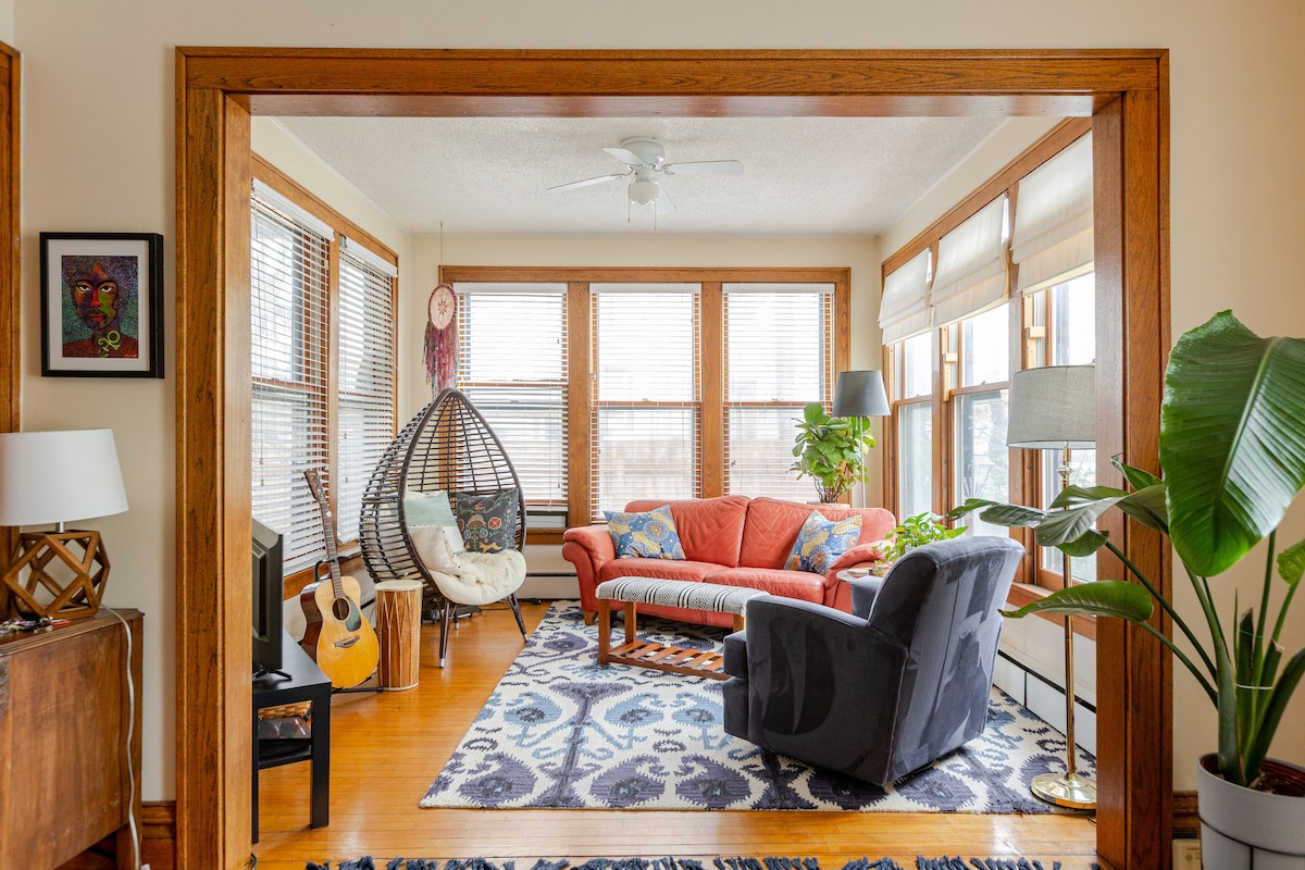 A sunlit living area features a cozy red sofa, a blue armchair, and a unique hanging chair. Natural light floods in through multiple windows, illuminating the wooden trim and vibrant area rug. Plants and art integrate warmth into the inviting space.