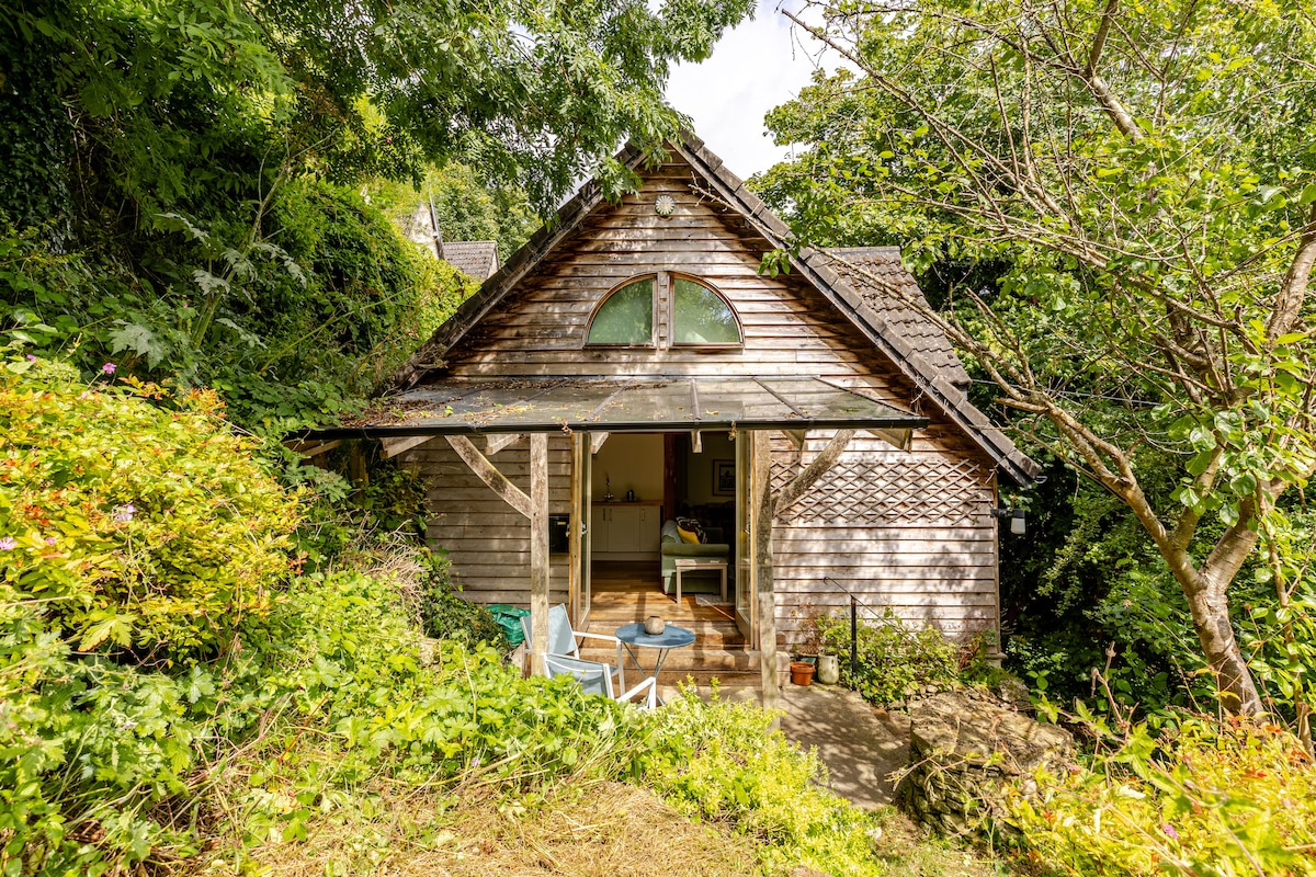 A wooden guest house is surrounded by lush greenery, with a large, sloped roof and arched windows. The entrance is framed by a covered porch, featuring two light blue chairs. The natural landscape enhances the private and serene setting.