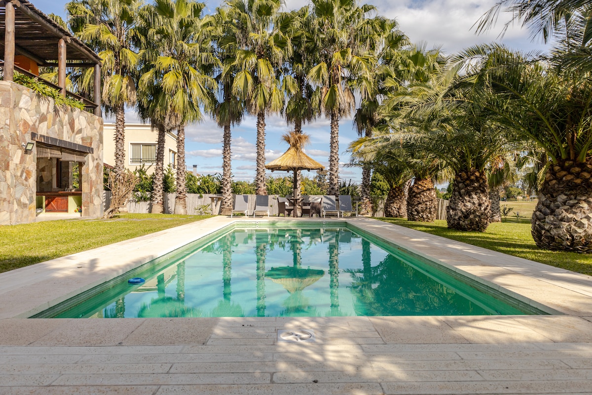 A private swimming pool is surrounded by palm trees and lush greenery. Comfortable lounge chairs are positioned nearby under a thatched-roof gazebo, creating a relaxed outdoor setting. The clear water of the pool reflects the sky, inviting moments of leisure.
