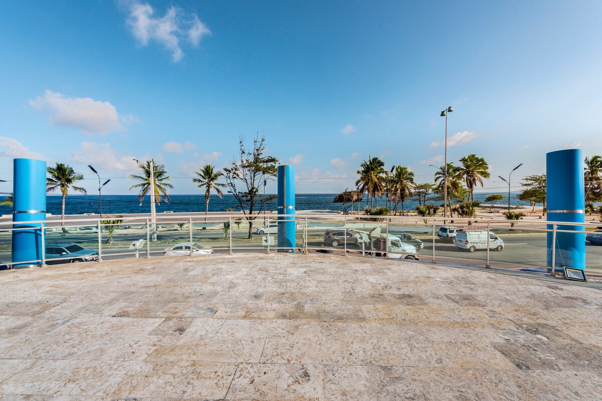A spacious balcony offers views of a serene ocean and palm trees, with a clear blue sky reflecting the coastal ambiance. The area is paved with light-colored stone, providing an ideal setting for relaxation.