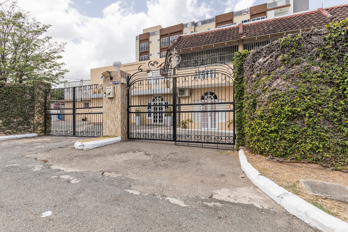 The exterior view of a well-maintained entrance gate features ornate ironwork and a stone pillar. The gate is framed by lush greenery, offering a sense of privacy and security. The surrounding area is paved, indicating careful landscaping and upkeep.