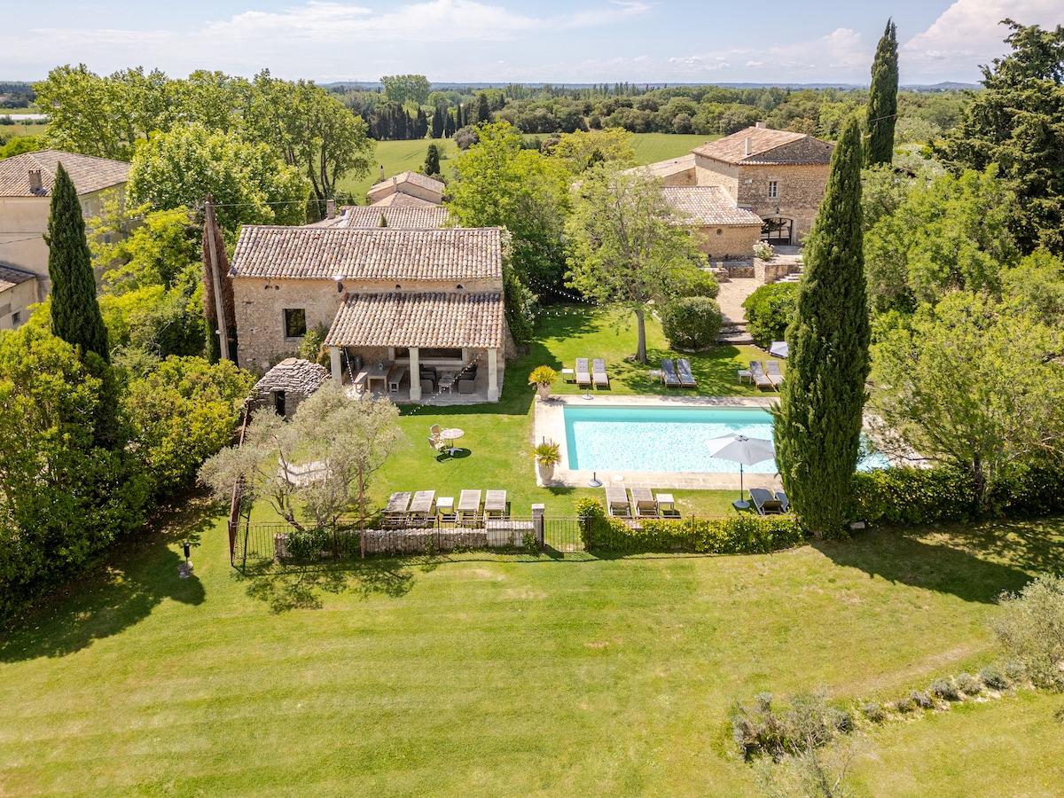 A spacious garden area surrounds a shared pool, complemented by sun loungers. The property features a charming house with distinctive roof tiles, bordered by lush greenery. Nearby, outdoor seating areas are arranged, creating inviting spaces for relaxation amidst a peaceful rural landscape.