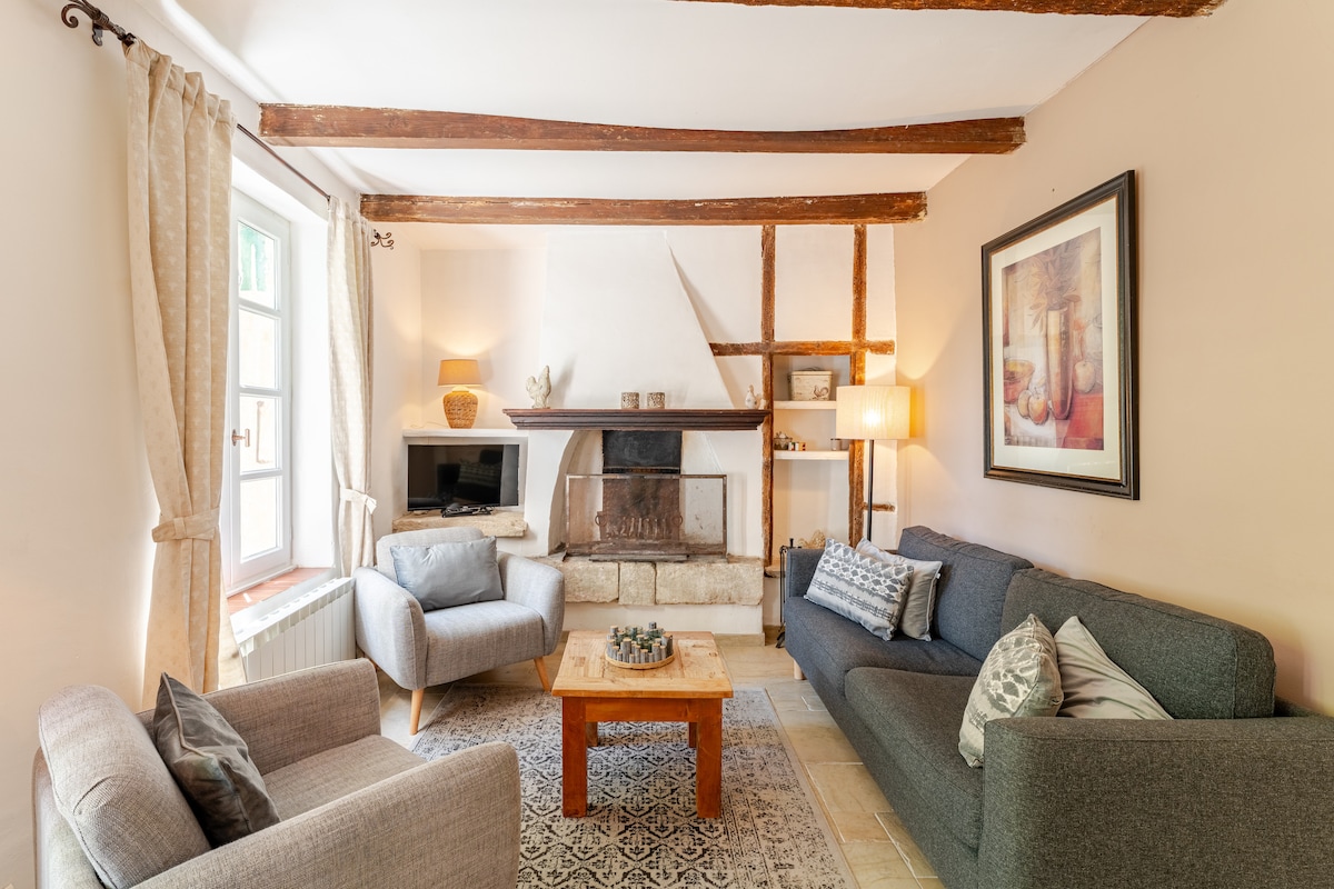 A cozy living area is presented, featuring two armchairs and a spacious sofa arranged around a wooden coffee table. A stone fireplace serves as a focal point against the backdrop of wooden beams. Natural light filters through the window, enhancing the warm ambiance.