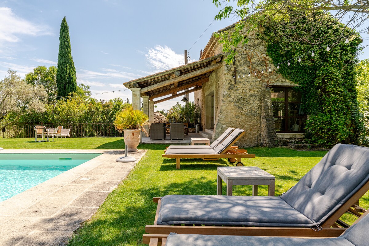 A tranquil outdoor space features comfortable loungers positioned on lush green grass near a shared pool. The rustic villa, adorned with climbing greenery, provides a charming backdrop under a clear blue sky, inviting relaxation in a serene setting.