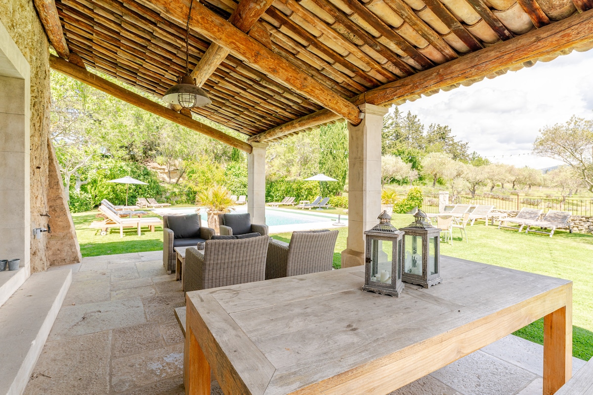 An outdoor seating area is shaded by a rustic roof with wooden beams. A wooden table is positioned in front, accompanied by decorative lanterns. In the background, a shared pool is visible, surrounded by sun loungers and lush gardens.