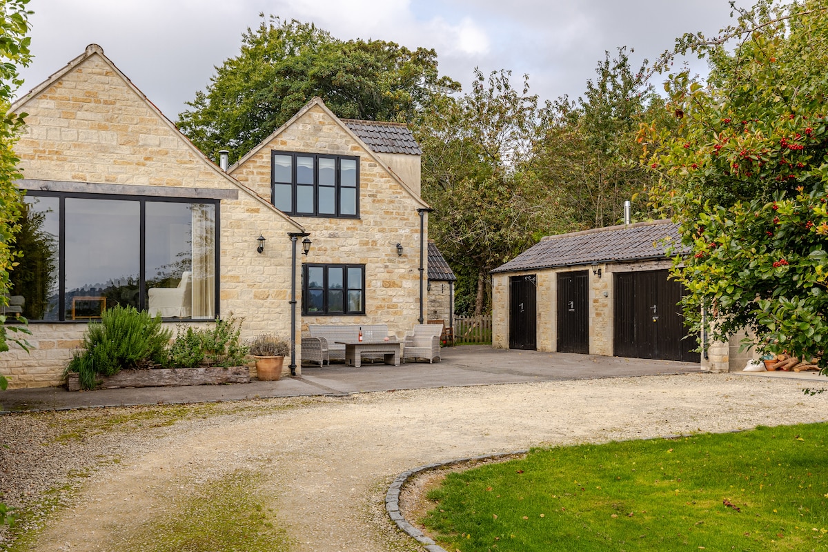 A charming stone home with large windows and a welcoming entrance is situated alongside a gravel driveway. The landscape features well-maintained greenery and a separate outbuilding. Outdoor seating is arranged for relaxation, creating an inviting exterior space for guests.