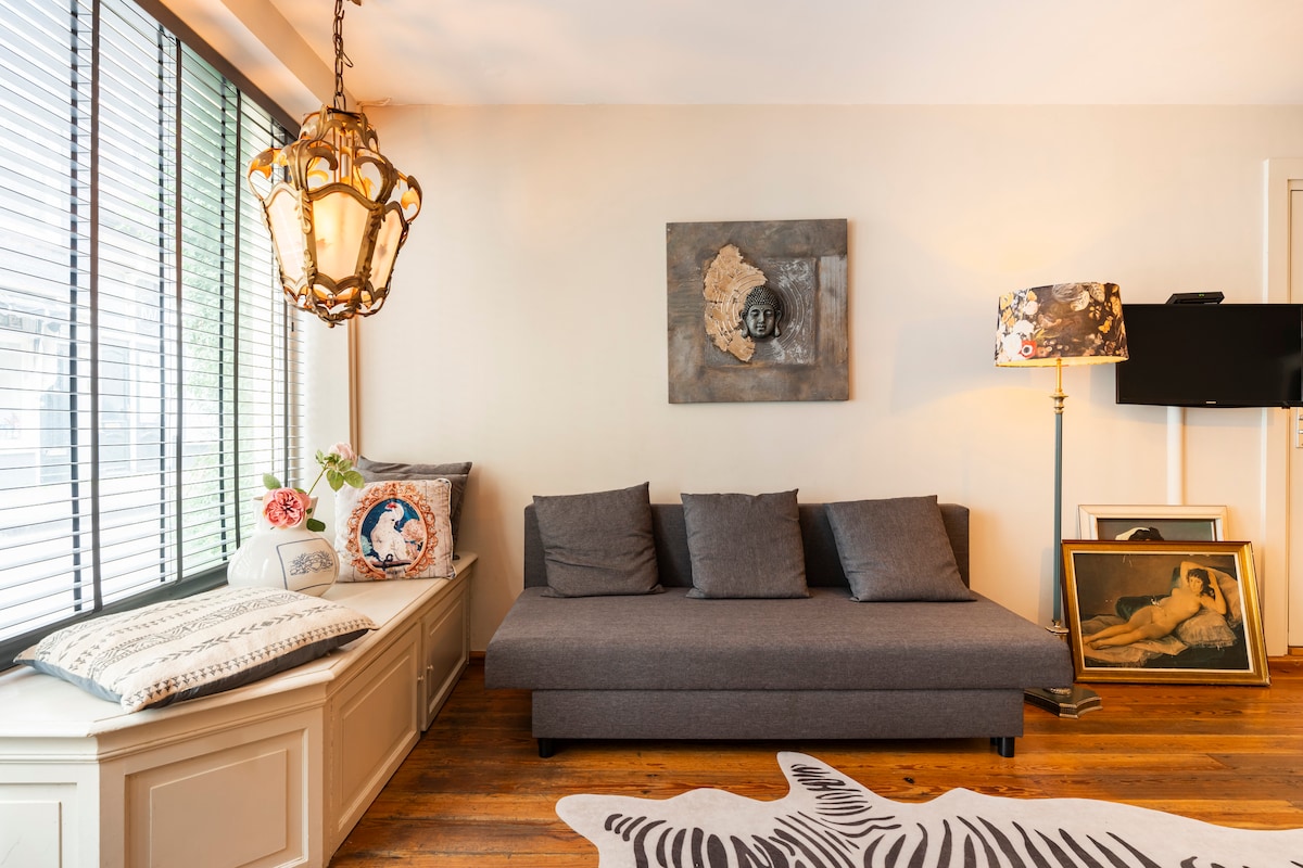 A cozy seating area features a gray fold-out sofa accompanied by a decorative lamp and artwork. Natural light floods in through the large, vertical windows, highlighting the wood floors and modern decor. An inviting atmosphere is created by the thoughtful arrangement of furnishings.