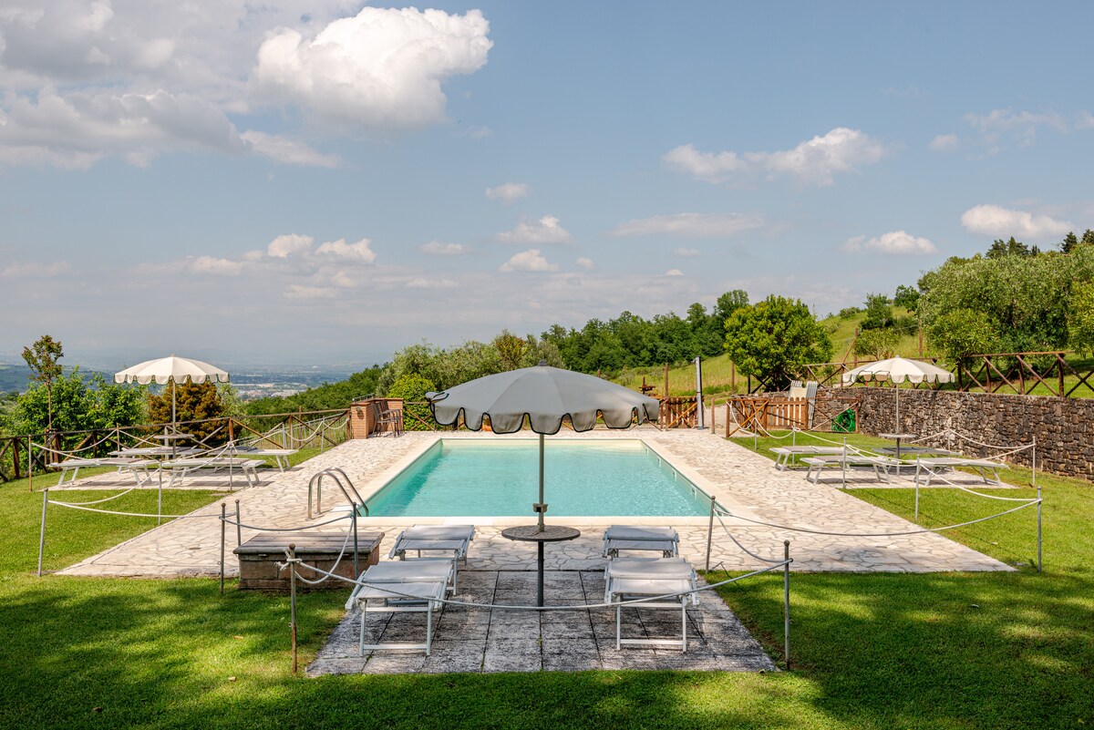 An outdoor swimming pool is surrounded by a stone patio, equipped with several lounge chairs for relaxation. An umbrella provides shade over a small table. Lush green hills in the background emphasize the serene environment.