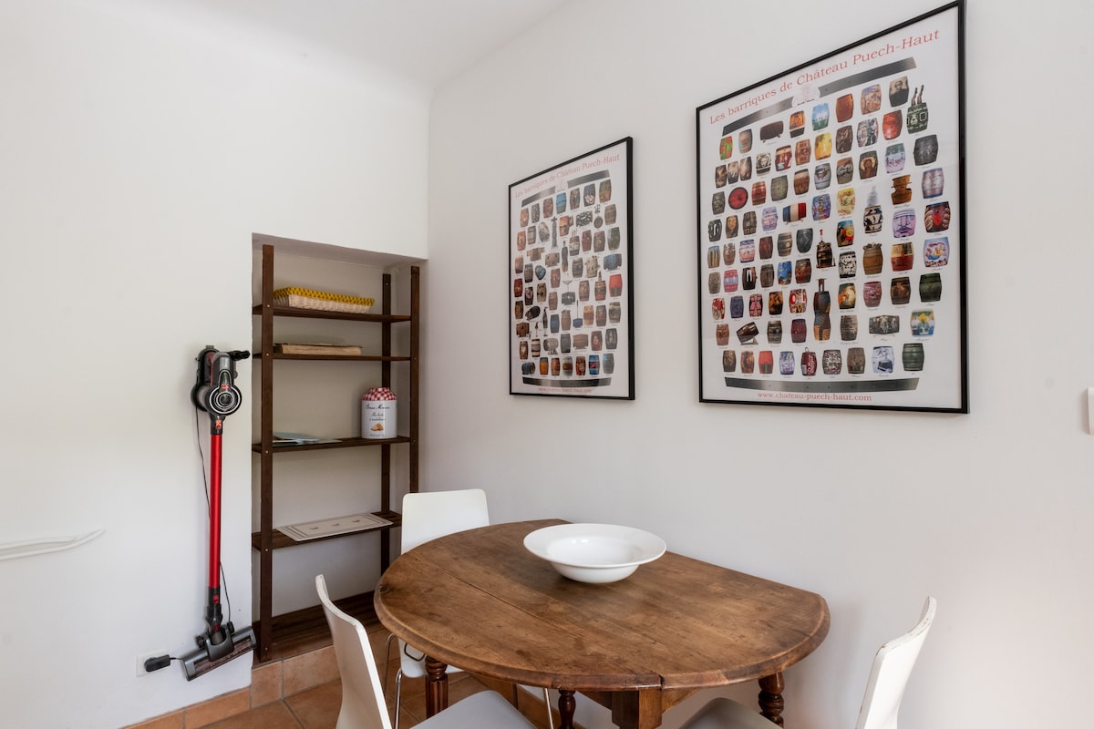 A simple dining area features a round wooden table accompanied by white chairs. On one wall, two framed collections of colorful images are displayed, while a shelf with various items is positioned in the corner. The space is well-lit with a neutral color palette.