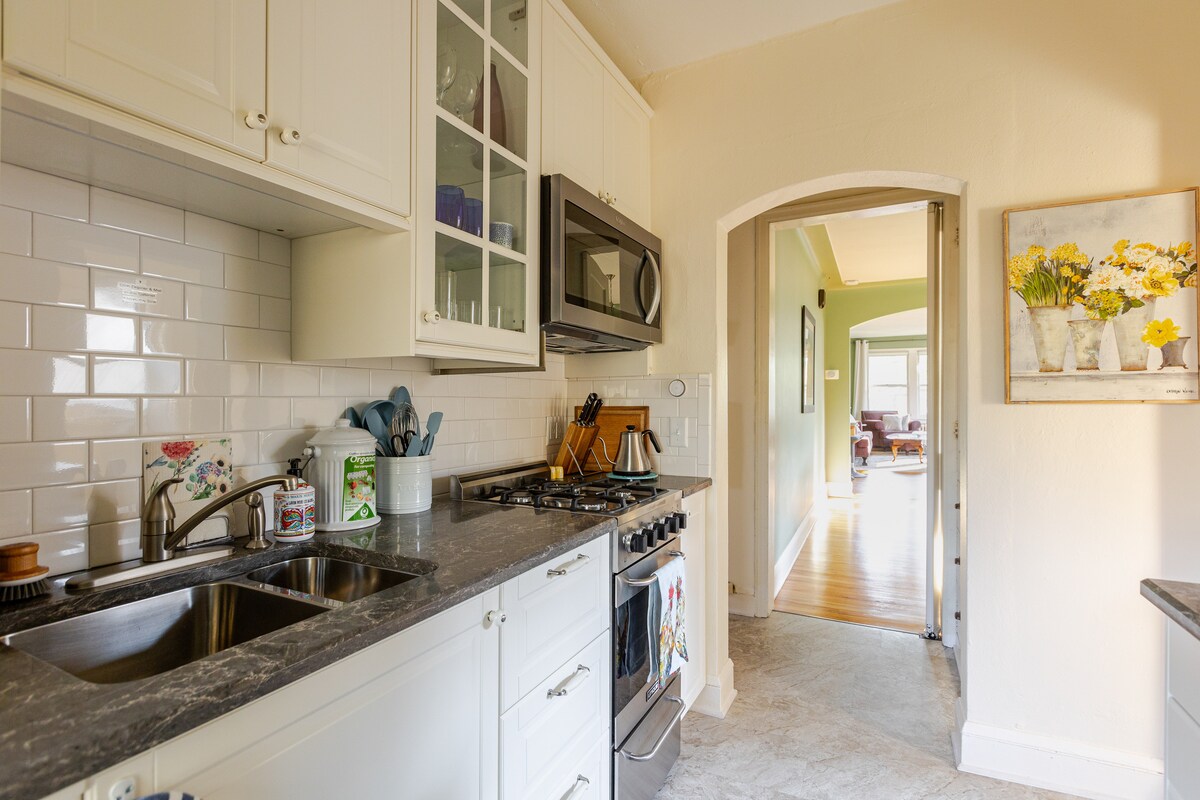 The kitchen is equipped with modern appliances including a microwave and stove. White cabinetry and a dark countertop create a clean aesthetic. A sink is positioned beneath a window, allowing natural light to brighten the space. An arched doorway leads to the living area.
